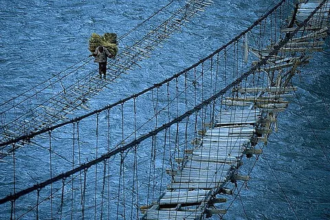 September 1991, Hunza River, Gojal, Pakistan --- A sure-footed Hunzakut with a load on his back walks across the new Hussaini bridge built to carry villagers and livestock over the Hunza River hanging beside the old bridge blown apart by winter winds, Gojal, Pakistan. --- Image by © Jonathan Blair/CORBIS
